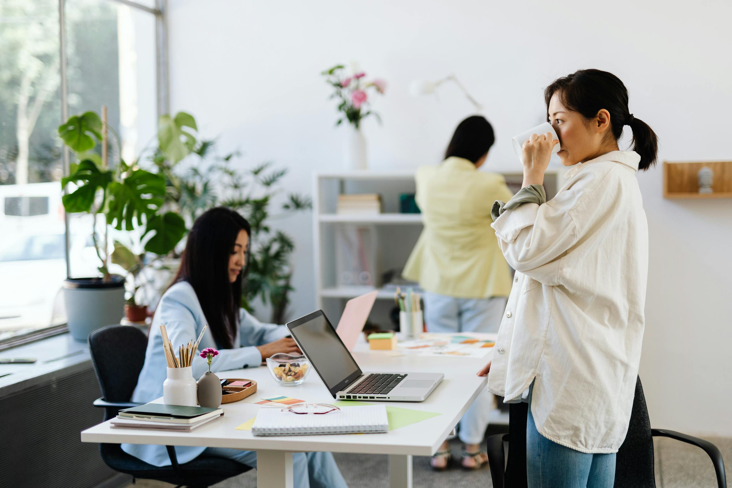 Three women in a modern office, working and interacting with laptops and plants around.