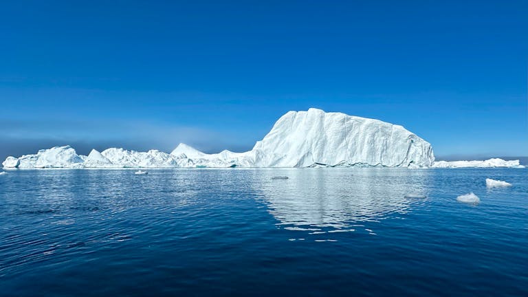 Beautiful iceberg reflecting in the calm waters of Greenland under a clear blue sky.